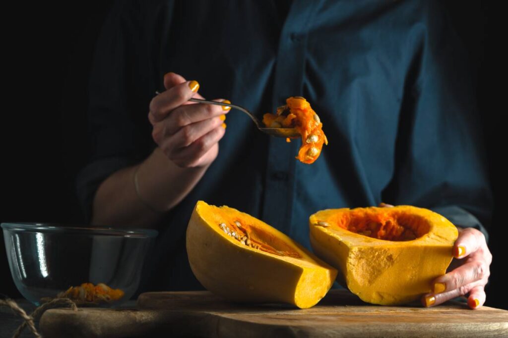 Spoon with seeds in a hand, pumpkin halves on a table on a blue background horizontal